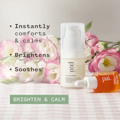 An arrangement of two skincare products from the brand pai, one in a white bottle with a pump and the other in a small orange dropper bottle, set among pink and cream flowers on a checkered surface, with text that reads Instantly comforts & calms, Brightens, Soothes, and Brighten & Calm.