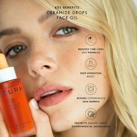 Image 1 - A close-up of a woman with blonde hair holding a serum bottle labeled Dr. Barbara Sturm Ceramide Drops Face Oil, with benefits outlined next to her: reduces fine lines and wrinkles, deep hydration boost, repairs compromised skin barrier, protects against harsh environmental aggressors.
Image 2 - An image showcasing two glass dropper bottles with ceramide face oil, highlighting skin benefit percentages for 1 DAY and 28 DAY, stating increases in skin barrier boost, hydration, and radiance, with the text CERAMIDE DROPS FACE OIL YOUR JOURNEY TO A HEALTHIER SKIN BARRIER IN ONLY 28 DAYS.
Image 3 - Image of a bottle of Dr. Baretts Ceramide Drops Face Oil with an orange liquid inside, accompanied by sections labeled Skin Identical Ceramides, Precious Oils, Astaxanthin, and PhytoSqualane detailing their benefits for skincare.
Image 4 - Image of a clear dropper filled with orange ceramide drops, positioned above a smooth white surface, with text stating Ceramide Drops Face Oil, 98% AGREE, and checkmarks next to claims about skin texture and protection.
Image 5 - A bottle of Dr. Barbara Sturm Ceramide Drops Face Oil is centered in the image, surrounded by orange-hued liquid droplets on a gradient orange background, with visible text that reads NEW CERAMIDE DROPS FACE OIL and A SKIN BARRIER BOOST WITH EVERY DROP.
Image 6 - The image features various skincare products, including jars labeled FACE CREAM, SUPER ANTI-AGING FACE CREAM, and FACE CREAM RICH, with swirls of white and orange creams around them, and includes text that reads CERAMIDE DROPS FACE OIL and HOW TO USE: Add a few drops of the CERAMIDE DROPS FACE OIL to your favorite Dr. Barbara Sturm MOISTURIZER to boost skin barrier strength, improve hydration and promote elasticity.