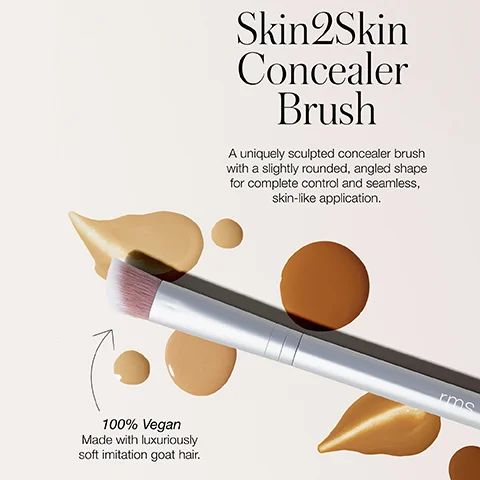 A silver-handled makeup brush with pink bristles is laid diagonally on a light background, surrounded by drops and smears of various shades of liquid foundation and concealer, along with the text Skin2Skin Concealer Brush, A uniquely sculpted concealer brush with a slightly rounded, angled shape for complete control and seamless, skin-like application, 100% Vegan Made with luxuriously soft imitation goat hair, and the brand name rms on the brush handle.