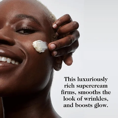 Image 2 - A person with dark skin smiles with a dollop of cream on their cheek and their hand near their temple, next to text that reads: This luxuriously rich supercream firms, smooths the look of wrinkles, and boosts glow.